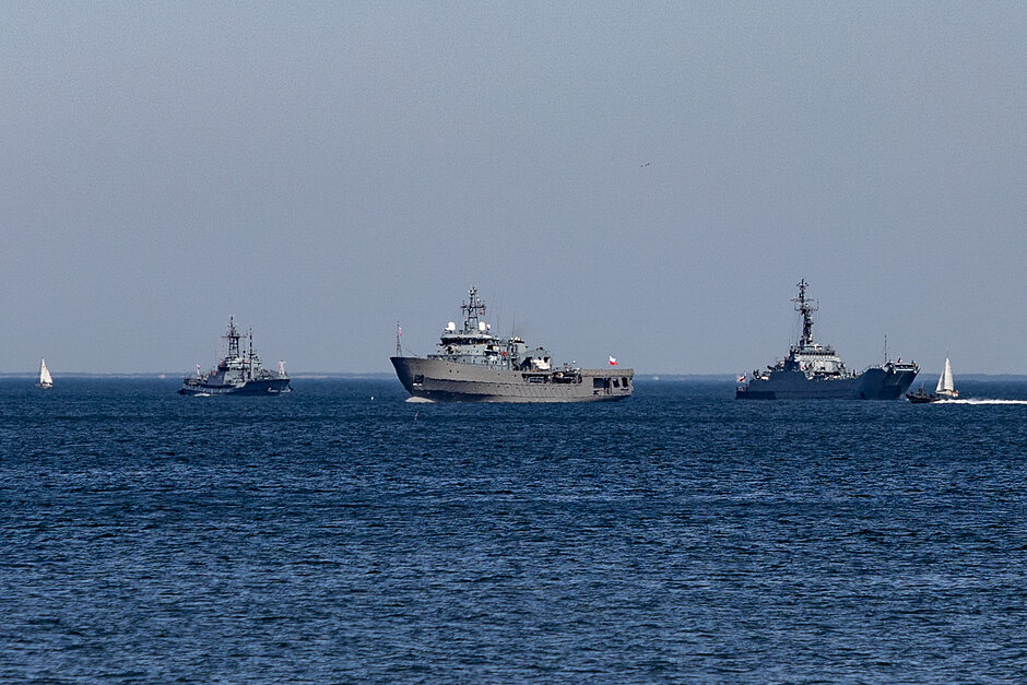 grupa trzech statków żeglujących po oceanie.Największy statek w centrum jest duży statek marynarki wojennej z szarym kadłubem i czerwoną flagą na górze.Wydaje się być sterowanym niszczycielem rakietowym, ponieważ na jego pokładzie widoczne jest kilka anten i innego sprzętu technicznego.Po lewej stronie znajduje się kolejny mniejszy statek, prawdopodobnie okręt wojenny lub holownik, z zawijającymi za nim żaglami.W tle można zobaczyć dwa mniejsze statki żeglujące obok większego statku.Obydwa statki są sylwetką na niebiesko, a woda jest spokojna.