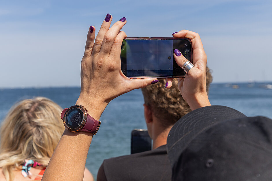 dłonie osoby trzymające smartfona do zrobienia zdjęcia trzymają telefon obiema rękami, z palcami lekko zakręconymi wokół krawędzi, mają fioletowy lakier do paznokci na paznokciach, w tle są inni ludzie siedząc na ławce, patrząc na wodę i niebo, na zegarku osoba na pierwszym planie nosi czarny kapelusz.
