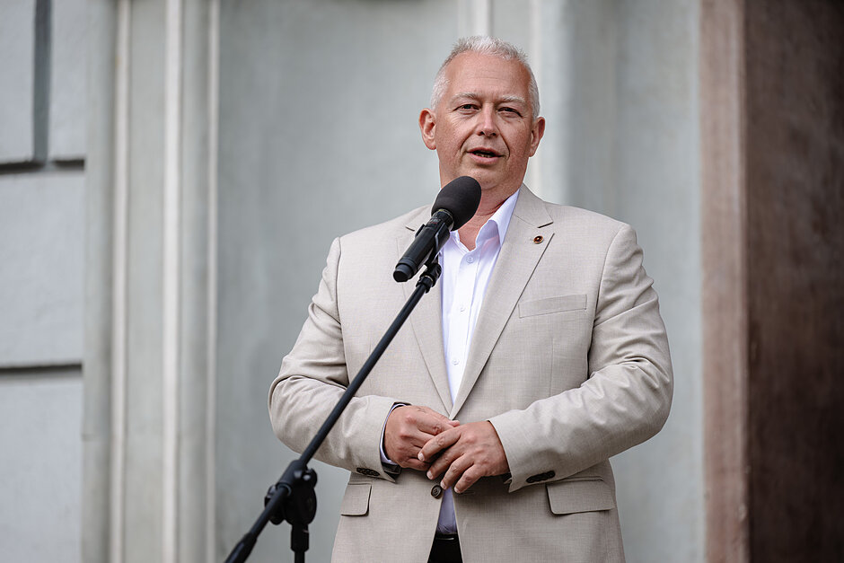 mężczyzna w średnim wieku stoi za mikrofonem i mówi do niego, ma na sobie lekki beżowy garnitur i białą koszulę, mężczyznę, który wydaje się być w późnych latach 60. lub na początku lat 70., z krótkimi białymi włosami, łysieje na górze głowy i ma krótko obcięte włosy, jego ręce są przywiązane do siebie i spogląda bezpośrednio na kamerę z poważnym wyrazem twarzy, za nim jest szara ściana z kolumnami po obu stronach, wydaje mi się, że człowiek wypowiada przemówienie lub prezentację.