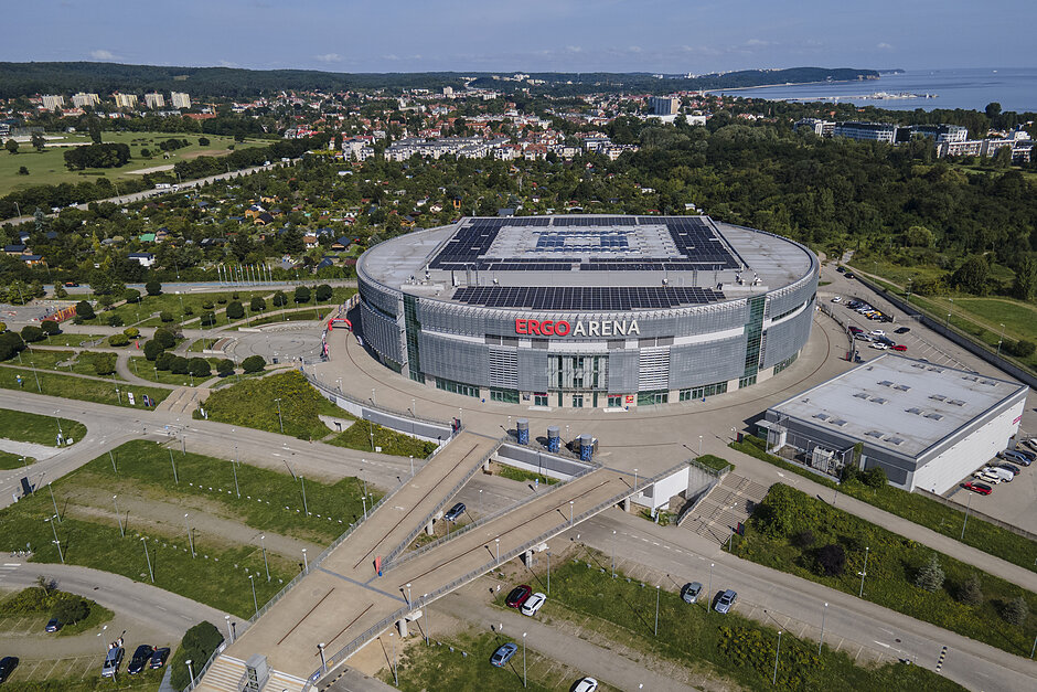 duży stadion sportowy z zakrzywionym dachem. Stadion znajduje się w zielonej okolicy z drzewami i zielenią wokół niego. W tle znajdują się kilka budynków i domów, a po prawej stronie obrazu widać zbiornik wody. W środku tego zdjęcia stadion ma czerwono-biały znak, który brzmi ERGO ARENA . Niebo jest niebieskie, i pogoda wydaje się słoneczna i przejrzysta. Na parkingu i kilku drogach prowadzących do areny znajduję się kilkanaście samochodów.