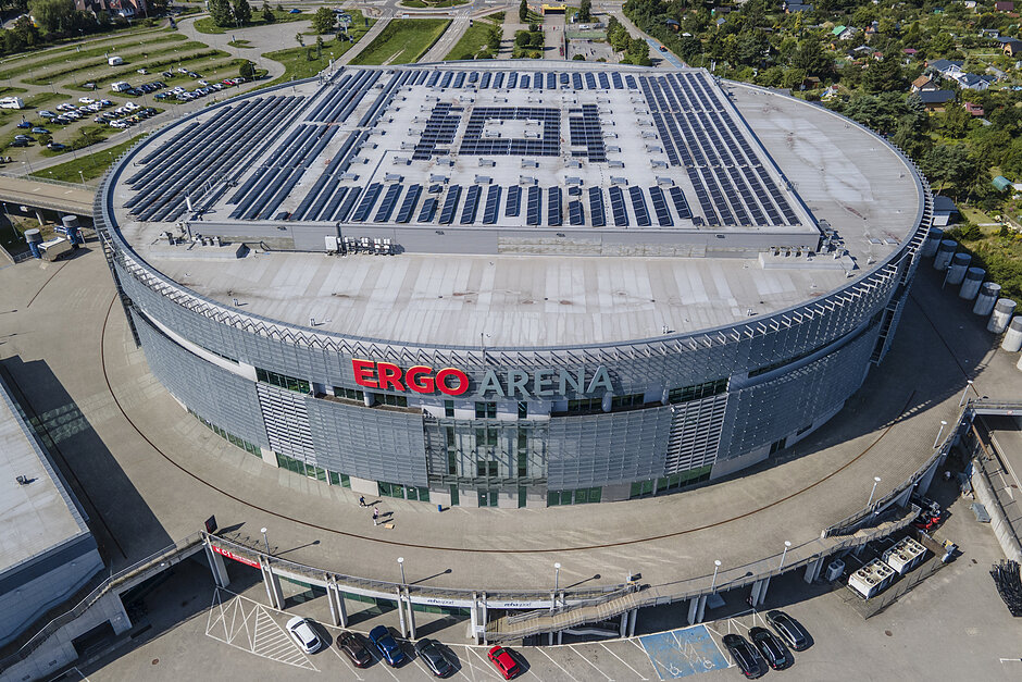 Ergo Arena, duża arena sportowa położona w przestrzeni miejskiej.Aerna jest okrągłym budynkiem z płaskim dachem i wieloma panelami słonecznymi pokrywającymi cały dach.Na przedniej fasadzie słowo ERGO  jest zapisane czerwonymi literami.Na parkingu pod areną zaparkowanych jest kilka samochodów i można zobaczyć kilka osób spacerujących.W tle widoczne są drzewa i inne budynki.