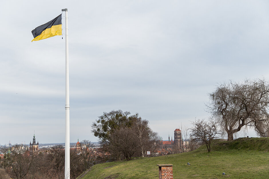 flaga z czarno-żółtą flagą latającą na wietrze. Flaga jest przymocowana do białego bieguna i znajduje się na trawnym wzgórzu. W tle widok na miasto z budynkami i drzewami. Po prawej stronie jest pochmurna, a ogólna atmosfera ponura.