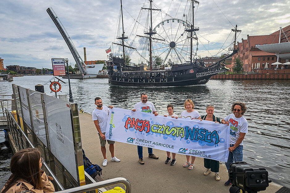Grupa osób stoi na brzegu rzeki w mieście i trzyma baner z napisem Akcja czysta Wisła .
