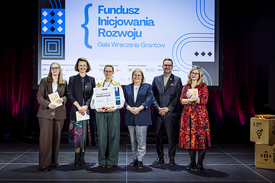Grupa kilku osób na scenie, pozują do zdjęcia z dyplomami 