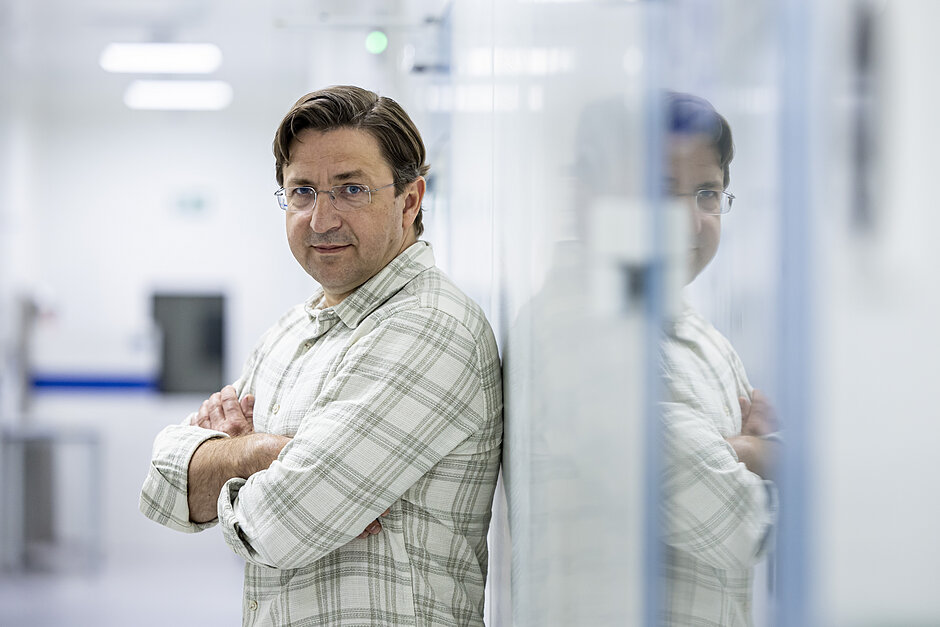  Stoi w laboratorium mężczyzna w średnim wieku, z krzyżowanymi rękami, nosi koszulę i okulary i ma poważne wyrażenie na twarzy, tło jest zamazane, ale wydaje się, że jest to czysta i nowoczesna przestrzeń z białymi ścianami i błękitną podłogą, po prawej stronie obrazu znajdują się szklane drzwi, a w tle można zobaczyć inną osobę.