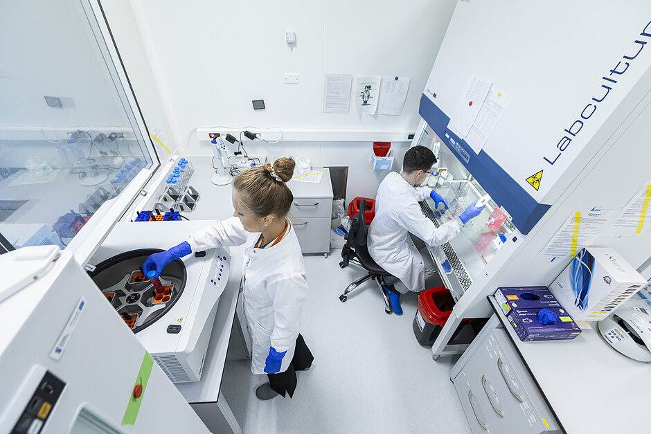  urządzenie laboratoryjne z dwiema osobami pracującymi w nim, oboje mają na sobie białe płaszcze laboratoryjne i niebieskie rękawice; osoba po lewej to młoda kobieta o blond włosach związanych w bułce i w ręku trzyma pipetę; stoi przed dużą maszyną z panelem sterowania i wydaje się pracować nad częścią sprzętu; po prawej stronie jest inna osoba siedząca przy biurku z rozproszonymi papierami i innym swym srzędziem naukowym; znajduje się również znak z napisem "Labkultura" oraz obudowa z różnymi instrumentami naukowymi i sprzęcie w tle.