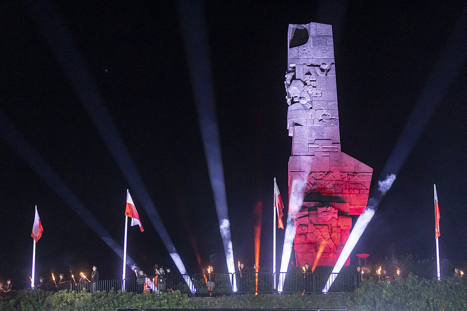 duży pomnik o czerwono-białym wyglądzie na nim. Pomnik wydaje się być wykonany z kamienia lub betonu i ma wysoki i prostokątny kształt. Jest oświetlony niebieskimi i czerwonymi światłami, tworząc dramatyczny efekt. Przed pomnikiem stoi kilka czerwonych flag z białymi biegunami i grupa ludzi stojących na nim, patrzących w górę na rzeźbę.