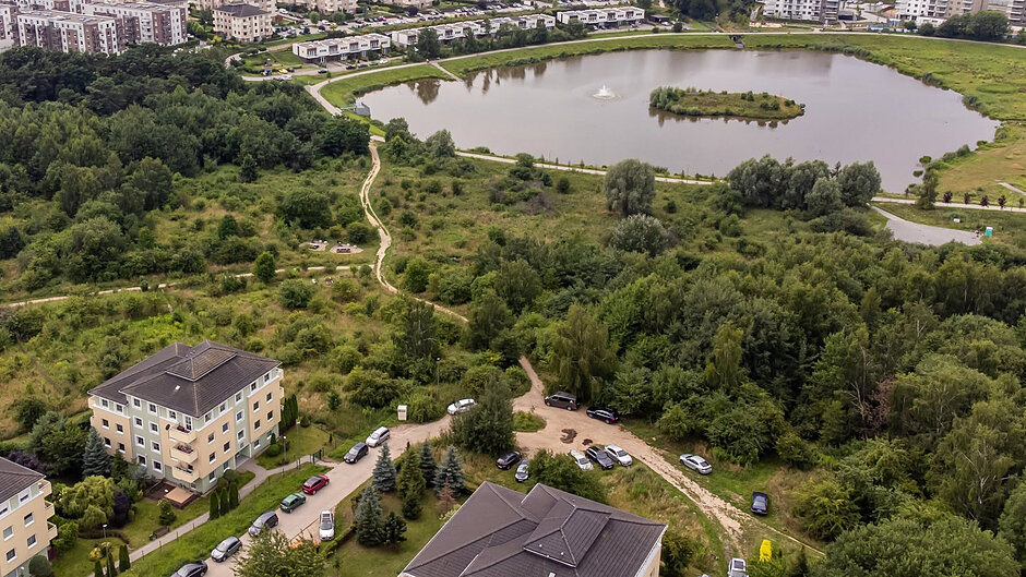 Zdjęcie z lotu ptaka, widok na zieloną przestrzeń ze stawem, wokół której stoją bloki mieszkalne.