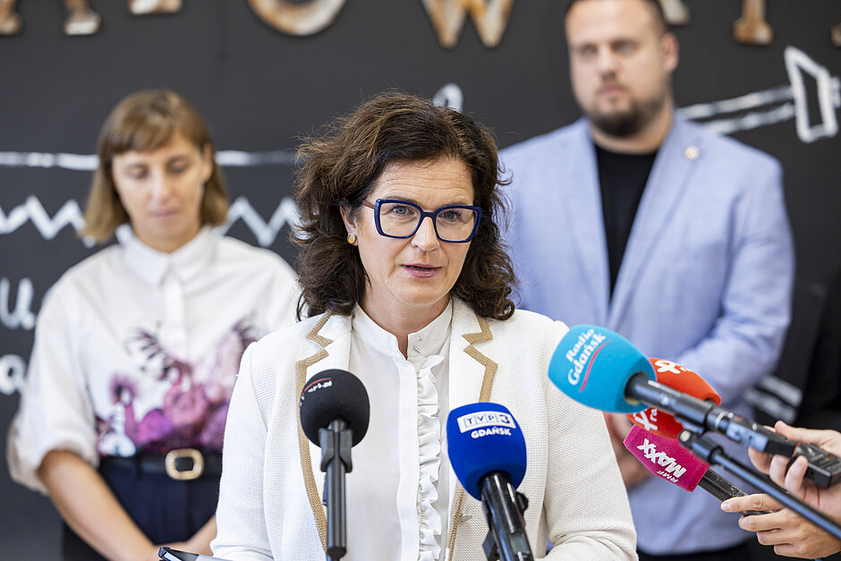 na zdjęciu prezydent gdańska mówi do mikrofonów dziennikarskich, ma kremową marynarkę