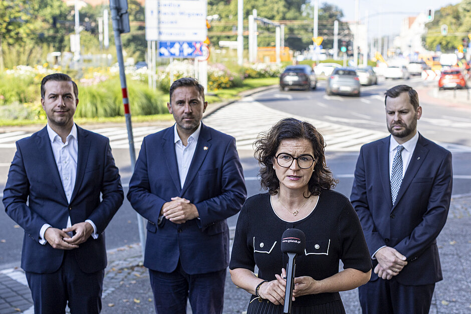 Prezydent Dulkiewicz podczas konferencji prasowej, stoi przy mikrofonie. Zaraz za prezydent stoją trzej inni uczestnicy konferencji