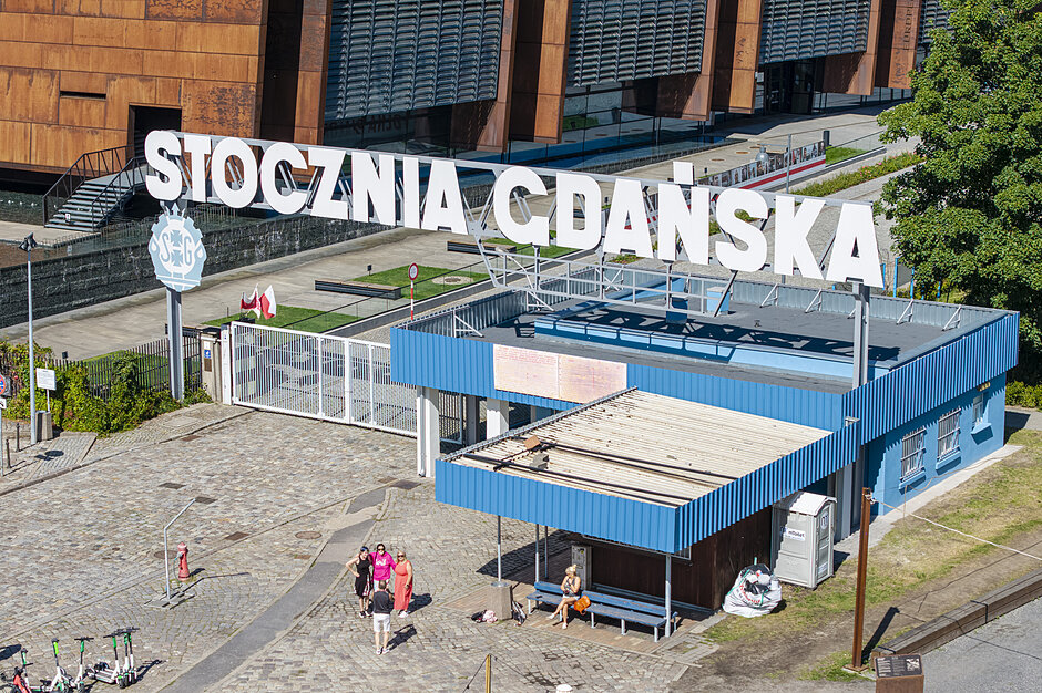  widok z powietrza niebieskiego budynku z napisem Stocznia Cdanskka  w białych literach. Budynek wydaje się być dworcem kolejowym lub dworca kolejowego, ponieważ nad wejściem znajduje się duży znak ze słowem  STOCZNIA CODANSKA  napisanym na nim. Kilka osób chodzi po budynkach, niektóre z nich siedzą na ławkach, a inne na chodniku. Po prawej stronie obrazu widoczne są drzewa i inne budynki.