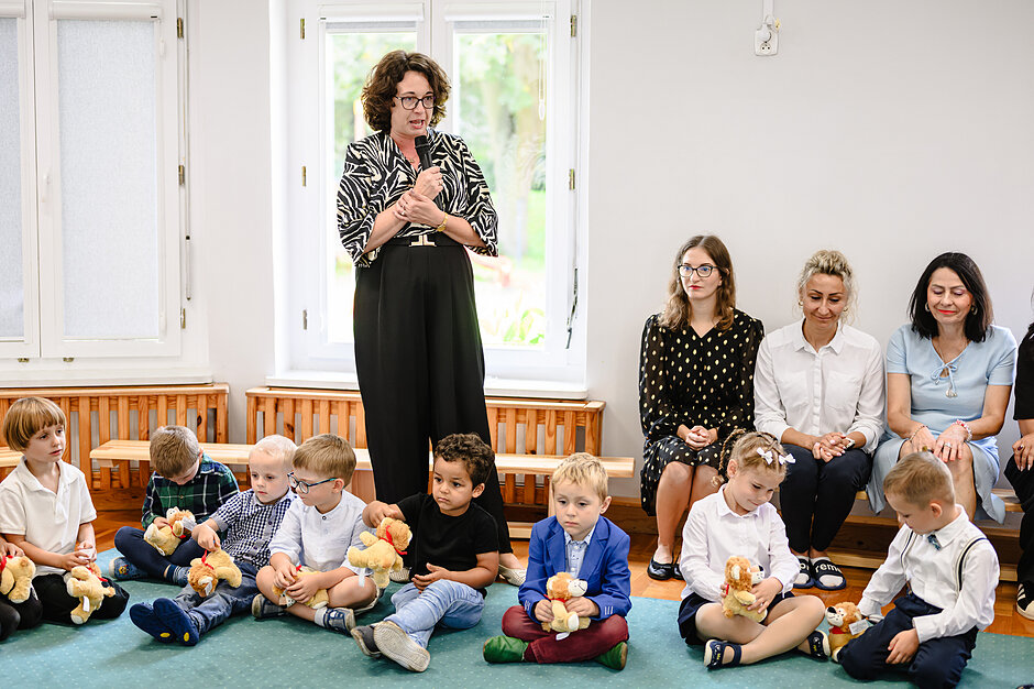 Rozpoczęcie roku szkolnego w przedszkolu nr 31. Dyrektor przemawia przez mikrofon. Obok niej siedzą dzieci z maskotkami. 