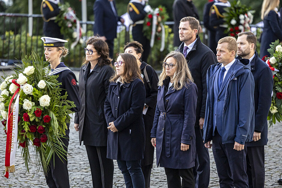 Grupa osób stoi za kobietą w marynarskim mundurze, która trzyma wieniec