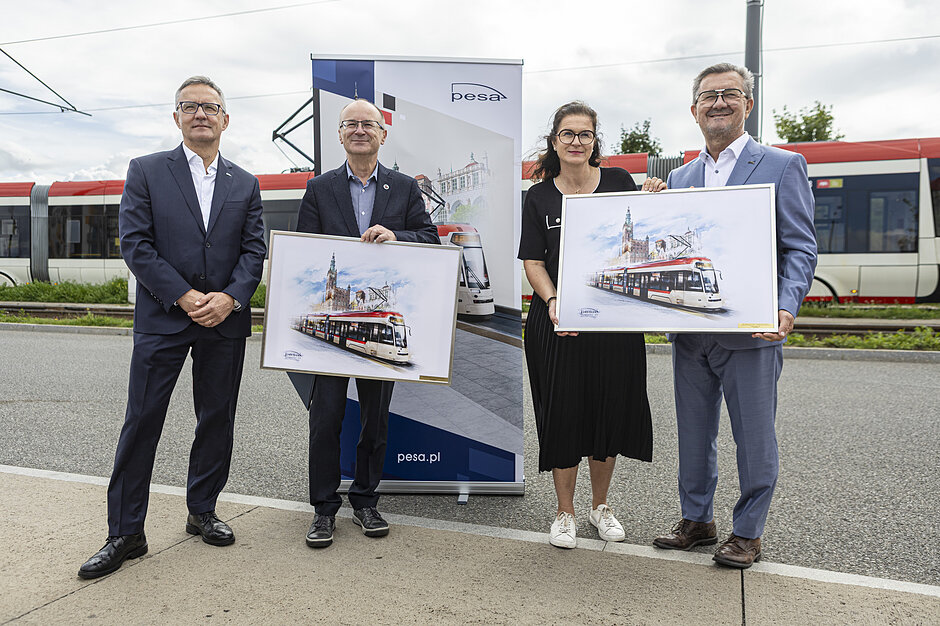 cztery wymienione osoby pozują trzymając dwie identyczne grafiki przedstawiające tramwaj