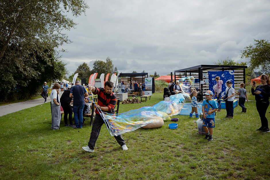 mężczyzna puszcza wielką bańkę, za nim pełno ludzi korzystających z różnych pikinikowych atrakcji, obok chłopie przyglądający się jego działaniom