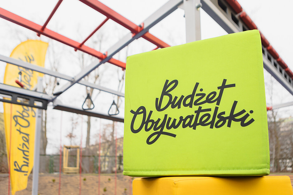 na zdjęciu zielona kostka z czarnym napisem budżet obywatelski, w tle urządzenie siłowni plenerowej