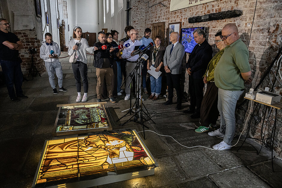 Kilka osób stoi wokół położonych na ziemi na podświetlonym stole witrażach w kościele.