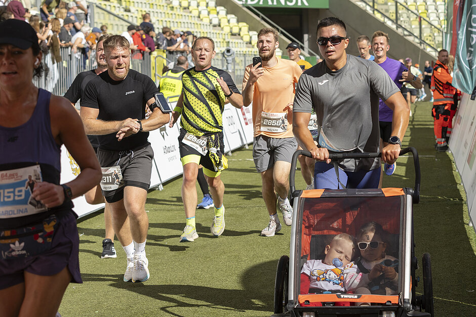 Biegacze biegną do mety na stadionie, wśród nich mężczyzna, który pcha wózek z dwójka dzieci.