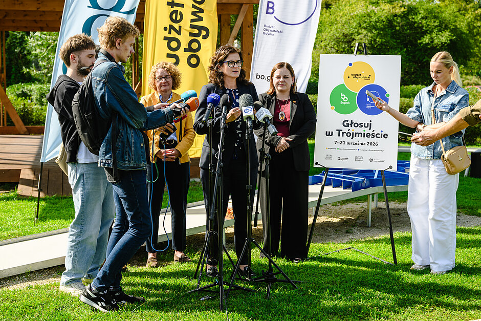 Trzy kobiety stoją w plenerze przed mikrofonami, obok stoją dziennikarze.