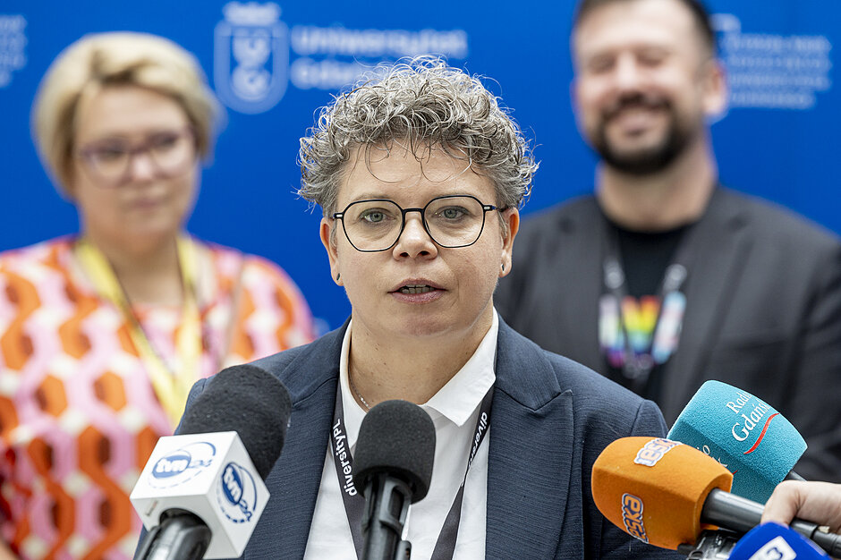 kobieta w średnim wieku mówi do mikrofonu, za nią stoją dwie osoby, których nie widać wyraźnie