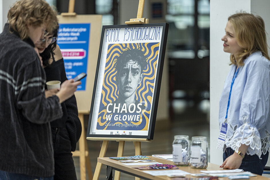 uśmiechnięta młoda kobieta patrzy na osobę, której twarzy nie widać w kadrze. między nimi plakat z napisem Pomoc psychiczna. chaos w głowie? 