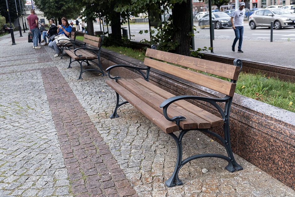 Wyremontowane ławki na skwerze w mieście, na niektórych siedzą ludzie.