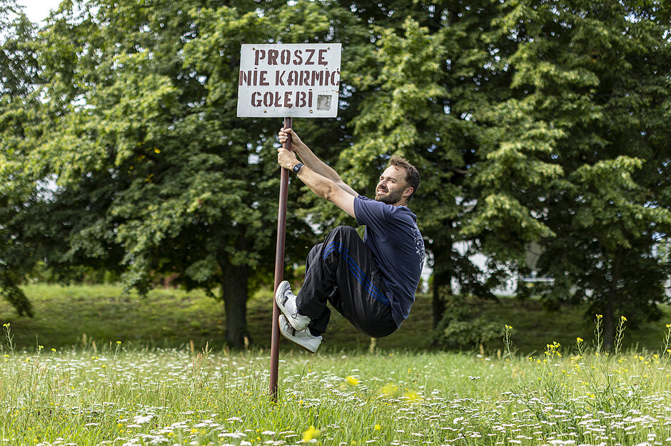 człowiek wiszący do góry nogami na słupie na polu wysokiej trawy i dzikich kwiatów. Ma na sobie niebieski t-shirt, czarne spodnie i białe trampki. Na spodzie znajduje się dołączony znak Prosze nie karmic golebi . W tle widoczne są drzewa i dom.
