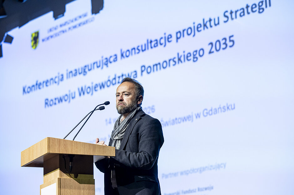mężczyzna stoi przy mównicy ,za nim ekran z napisem informującym o temacie konferencji