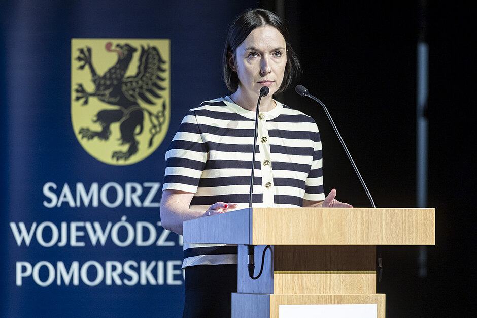 kobieta przy mównicy, za nią herb pomorza - czarny gryf na żółtym tle oraz napis samorząd województwa pomorskiego