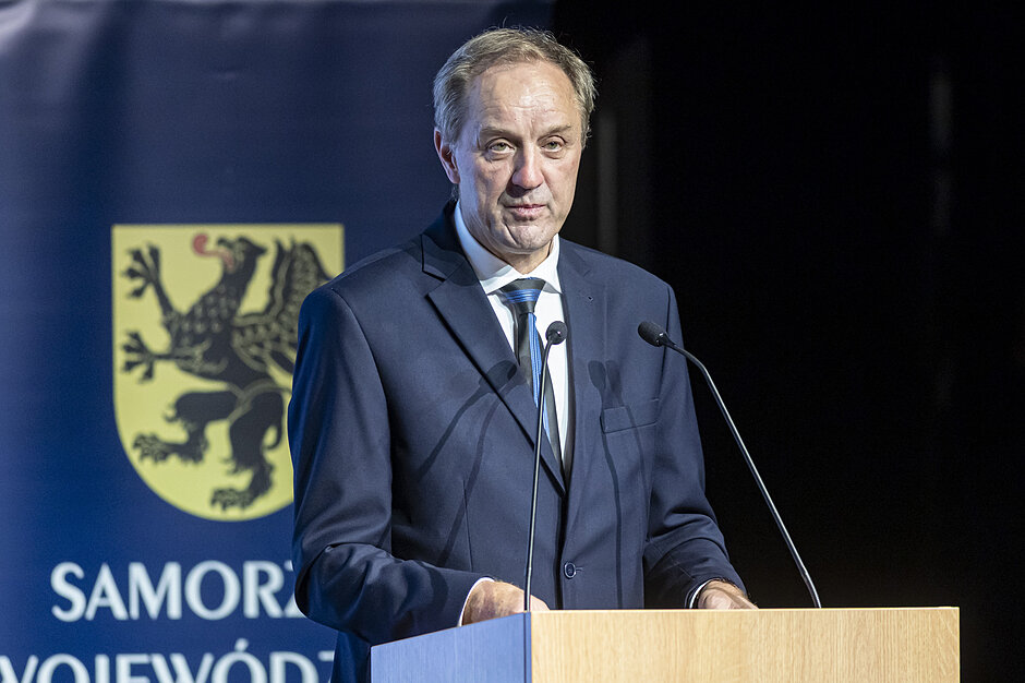 mężczyzna stoi przy mównicy, za nim herb pomorza i napis samorząd województwa
