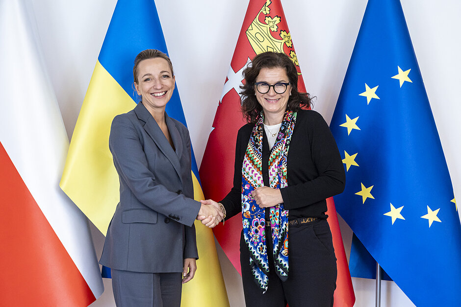 Dwie uśmiechnięte kobiety pozują do zdjęcia na tle flag Polski, Gdańska, Unii Europejskiej i Ukrainy