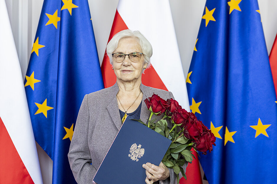 starsza kobieta stoi na tle flag polski i europy. z teczką z herbem polski i wielkim bukietem róż