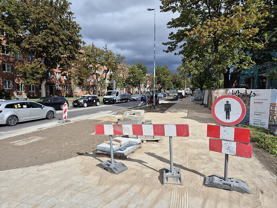 na zdjęciu znak drogowy biało czerwony z barierką uniemożliwiająca przejście przez remontowany chodnik