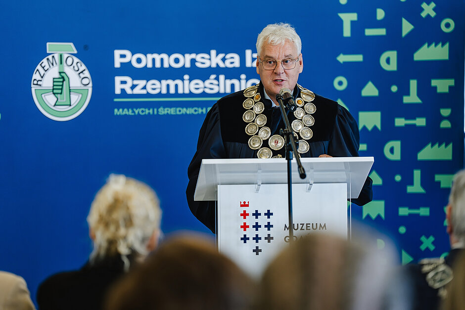 starszy mężczyzna w długiej granatowej szacie z ozdobnym łańcuchem przemawia przy pulpicie z napisem Muzeum Gdańska