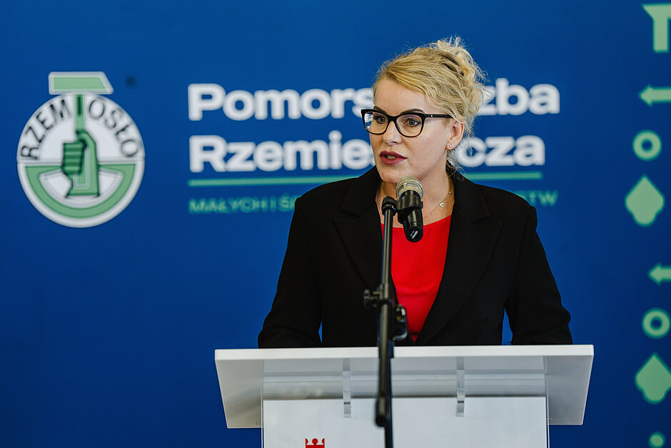 kobieta w średnim wieku przemawia przy pulpicie, za nią logo pomorskiej izby rzemiosł