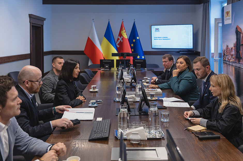 ludzie siedzą przy stole, przed nimi ekran prezentacyjny i flagi polski, ukrainy, gdańska i unii europesjkiej