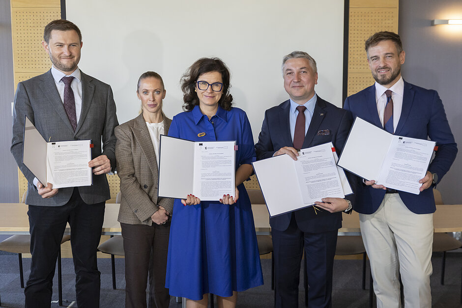 Na zdjęciu pozuje pięć osób w oficjalnych garniturach, trzech mężczyzn i dwie kobiety, pokazują podpisaną umowę