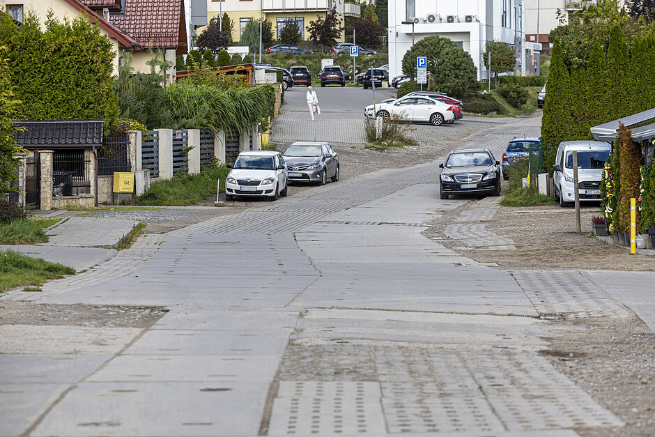 na zdjęciu ulica z tymczasowymi płytami betonowymi, po obu stronach zabudowa mieszkaniowa, widać też kilka zaparkowanych samochodów