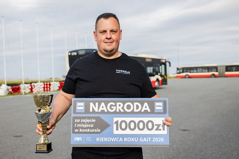 młody mężczyzna stoi z symbolicznym czekiem na 10 tysięcy złotych i pucharem w ręku, w tle autobus