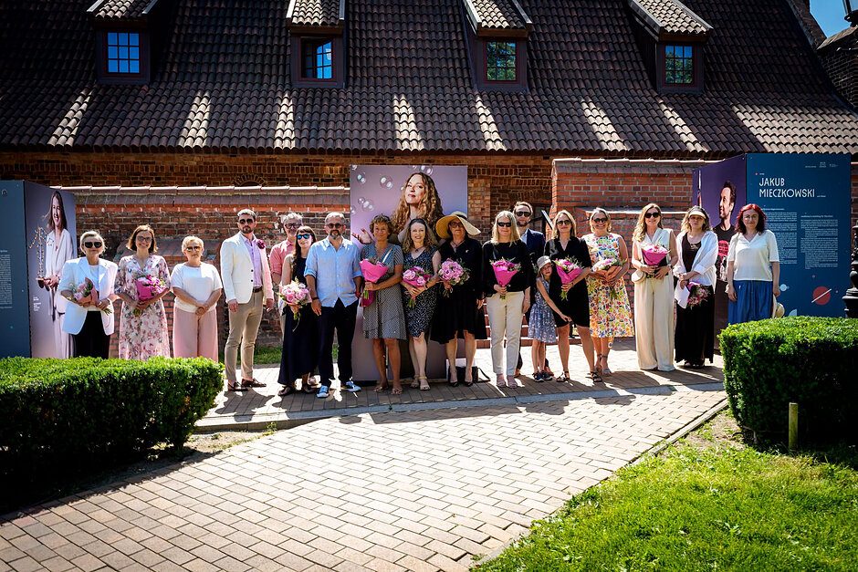 Liczna grupa osób pozuje do zbiorowej fotografii na tle wystawy plenerowej w mieście.
