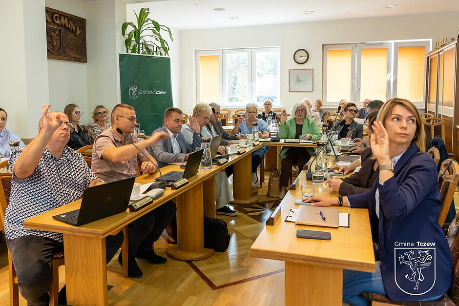 kilkanaście osób siedzi przy stole, podnosząc ręce do głosowania