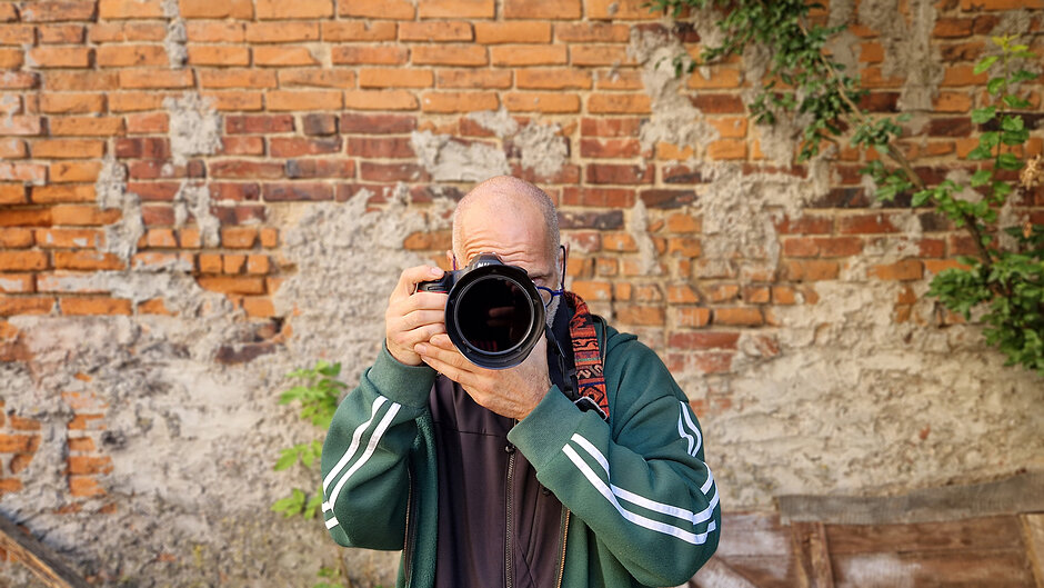 Fotograf na tle ceglanej ściany z wycelowanym obiektywem w naszą stronę