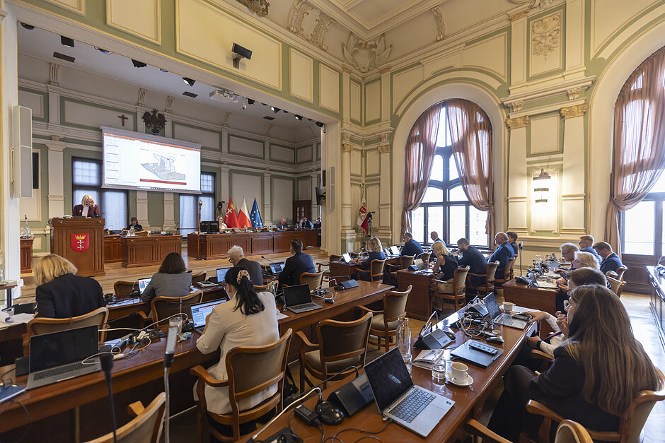 na zdjęciu sala obrad w zabytkowym wnętrzu, widać kilkanaście osób siedzących za stołami tyłem do fotografującego