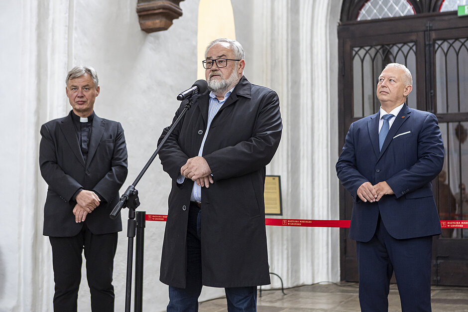 Trzy osoby stoją. Jedna z przodu przy mikrofonie