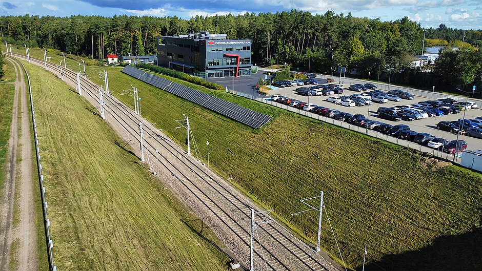 tory kolejowe, na skarpie zamontowana instalacja fotowoltaiczna, w tle budynek i parking pełen samochodów