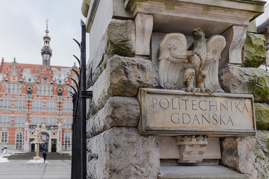 w tle duży gmach, z prawej, na pierwszym planie, furtka. Przy niej wykuty w piaskowcu orzeł, pod nim wyryty napis politechnika gdańska