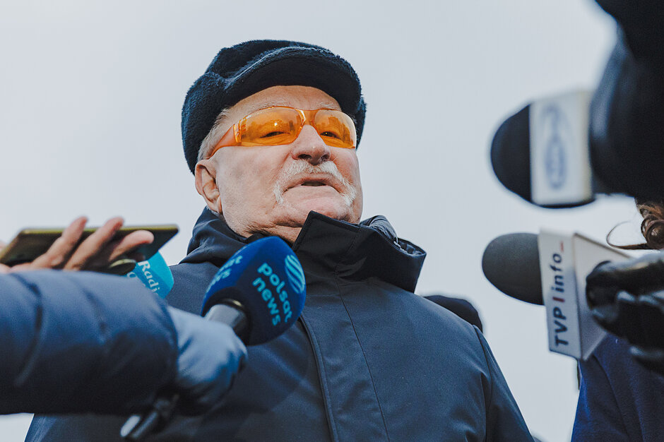 Lech Wałęsa w zimowym ubraniu i pomarańczowych okularach. Dookoła niego dużo dziennikarzy z mikrofonami. 