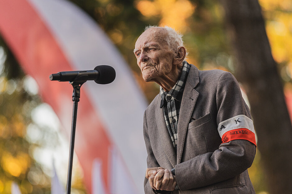 starszy mężczyzna w marynarce i z białoczerwoną przepaską na lewym ramieniu, mówi do mikrofonu
