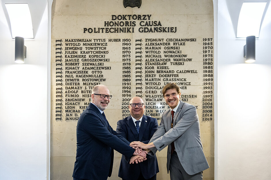 Trzech mężczyzn przed tablica z nazwiskami w hallu uczelni.