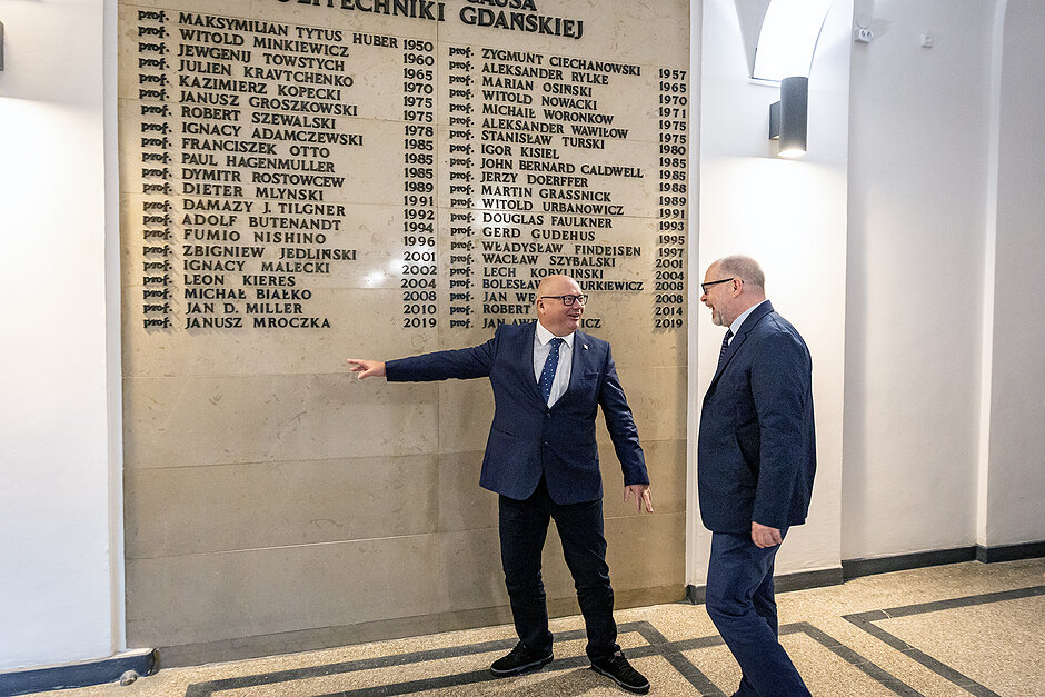 Dwóch mężczyzn w hallu uczelni przed tablica z nazwiskami.